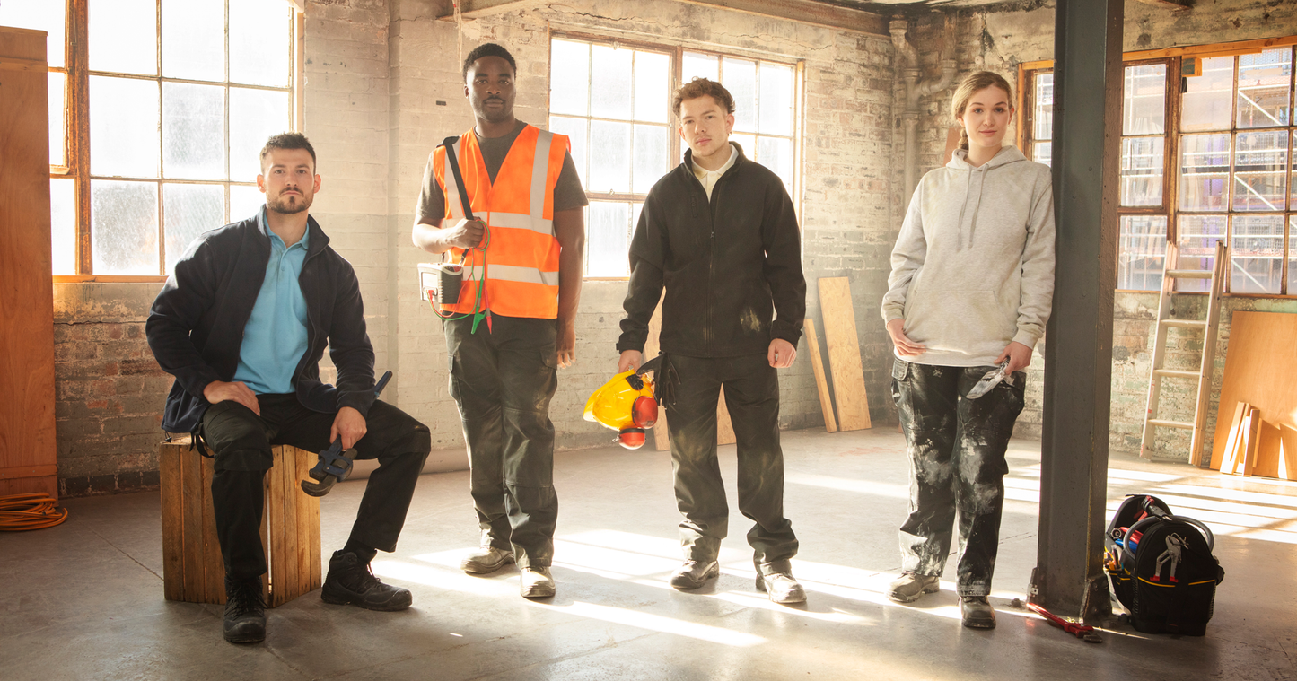 Four construction workers wearing Pro RTX workwear including polo shirts, hoodies, and safety gear, standing in a sunlit industrial workshop with tools and timber in the background.